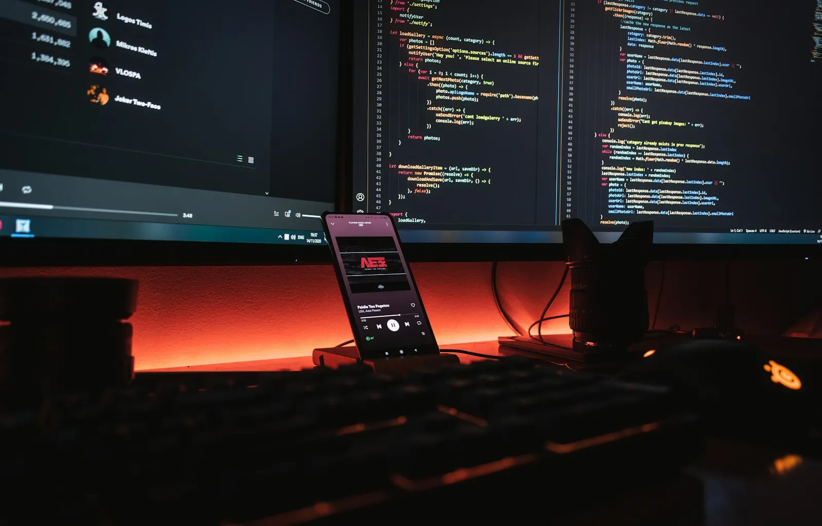 Monitores com código e um celular apoiado em uma mesa com luz alaranjada